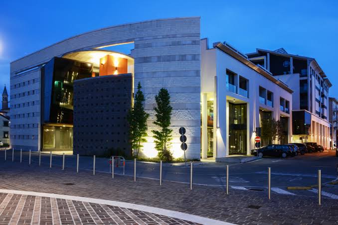 L’edificio del cinema all’imbrunire, visto dall’angolo della piazza: una facciata curva in pietra chiara con una grande vetrata illuminata di arancione, e alberi illuminati lungo il marciapiede.