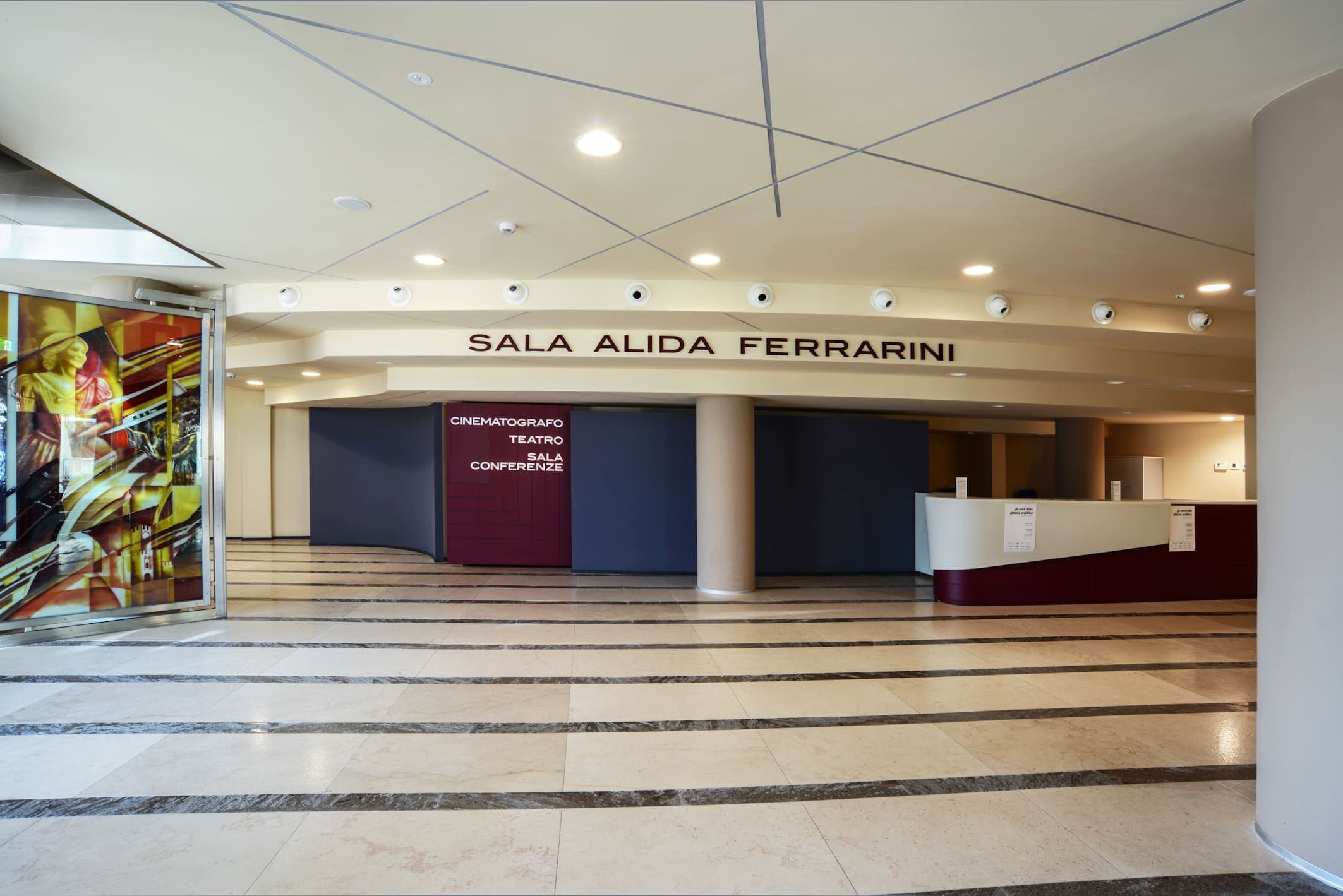 Il foyer, con il pavimento di marmo a fasce chiare e scure. Sopra le porte della sala la scritta SALA ALIDA FERRARINI; accanto, un pannello rosso con le parole cinematografo, teatro, sala conferenze. Sulla destra il bancone della biglietteria.