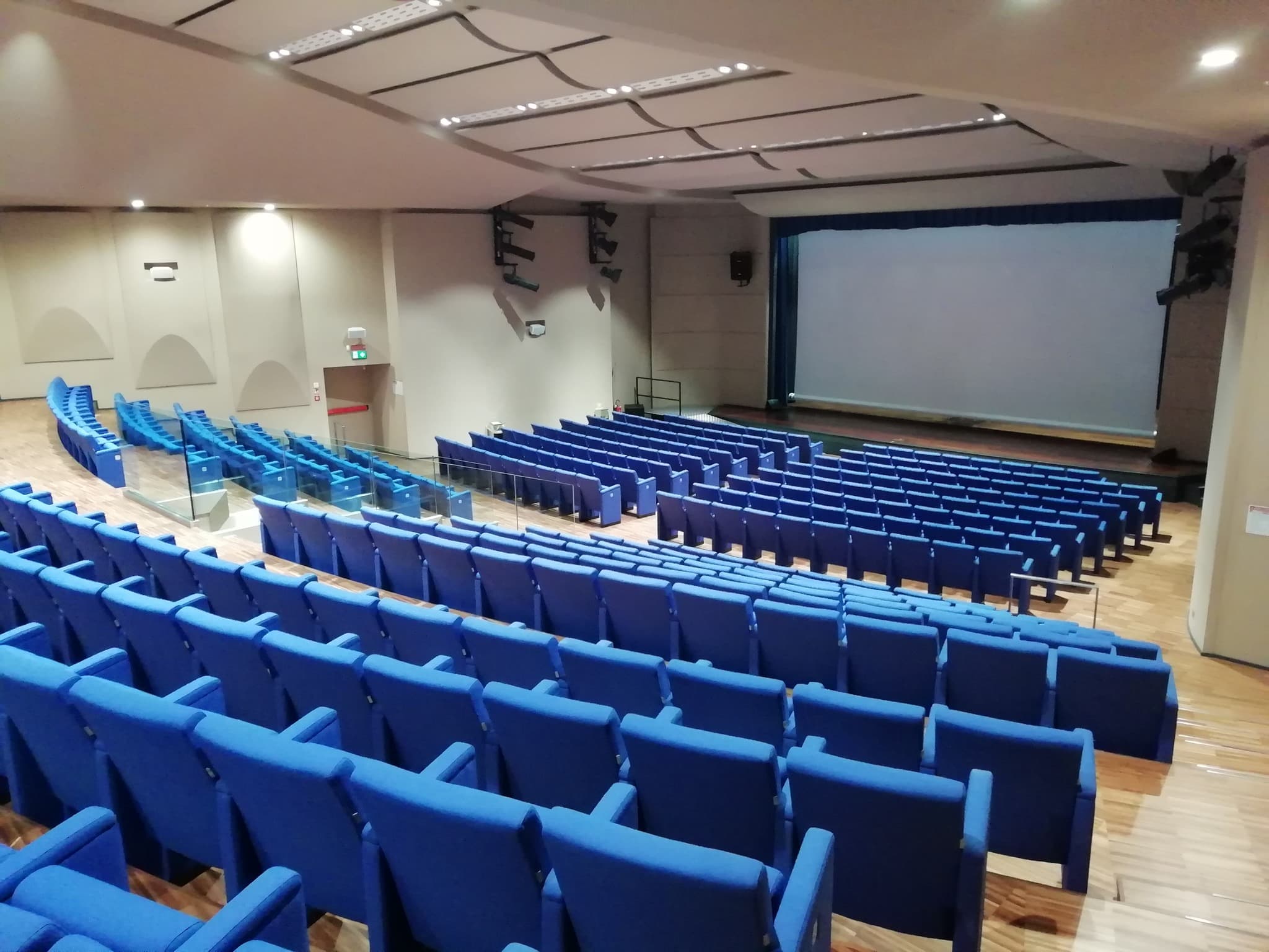 La platea vista dall’alto: lunghe file di poltrone blu su un pavimento in legno chiaro scendono a gradoni verso il palco. In fondo lo schermo bianco fra le tende blu, con i fari del teatro appesi ai lati.